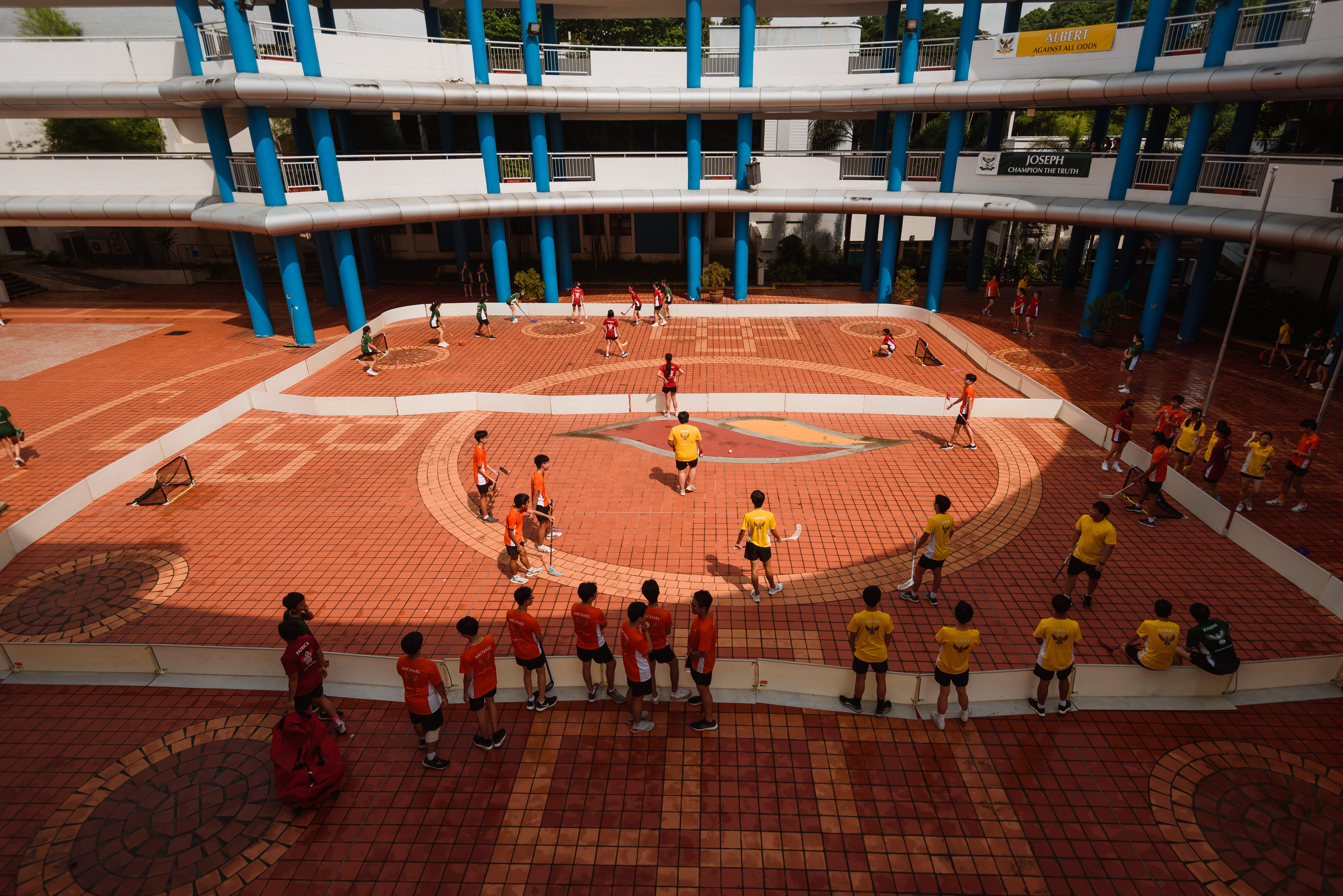 Students in orange and yellow play floorball on a brick court, watched by others, with blue columns behind.